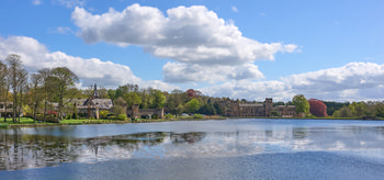 This landscape photograph captures the scenic view of Newstead Abbey lake and lodge from the fort on a bright spring morning. The calm water of the lake reflects the surrounding greenery and historic architecture, including the notable Newstead Abbey itself, which stands prominently in the background. The scene is defined by lush trees with fresh foliage lining the shore and a clear blue sky dotted with fluffy clouds. The combination of natural beauty and heritage buildings makes Newstead Abbey a significant landmark within the image, emphasizing the peaceful ambiance of the water and the picturesque estate in the heart of spring.